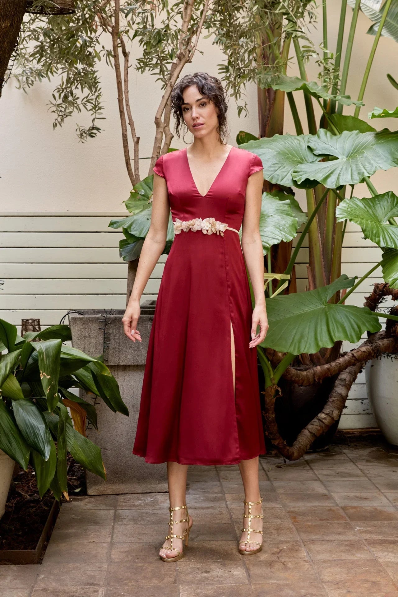 A woman stands in a patio garden wearing a deep red Multidress from la colección diseñada en España, featuring the CINTURÓN IBY floral belt and a thigh-high slit. She pairs it with strappy, studded sandals amid lush green plants.