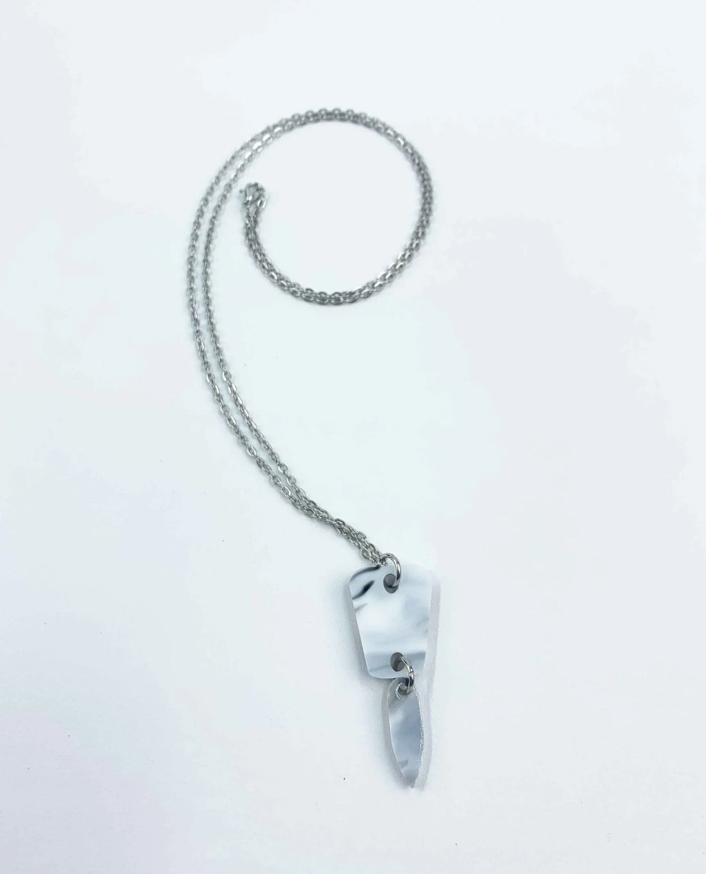 The Collar Minimalista de Cadena Plateada con Colgante Abstracto presenta dos piezas blancas y grises de forma irregular, tipo mármol, apiladas en un elegante diseño artesanal sobre fondo blanco.