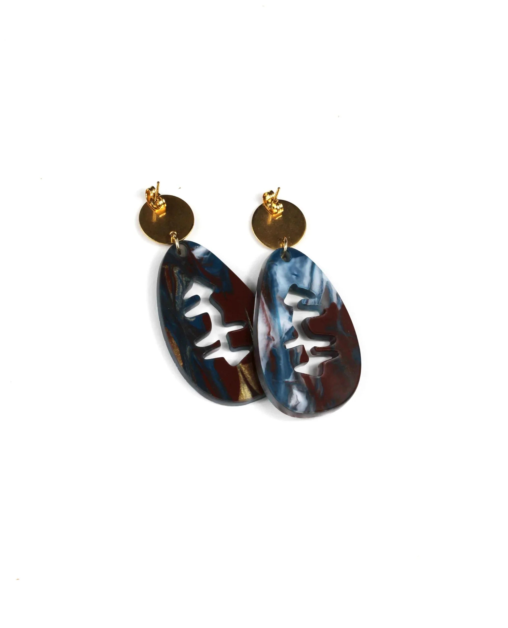 Two unique dangling earrings—Pendientes mármol marrón, azul, blanco y dorado—feature round gold studs and oval marbled blue and white pendants with abstract cutouts, set against a plain white background.