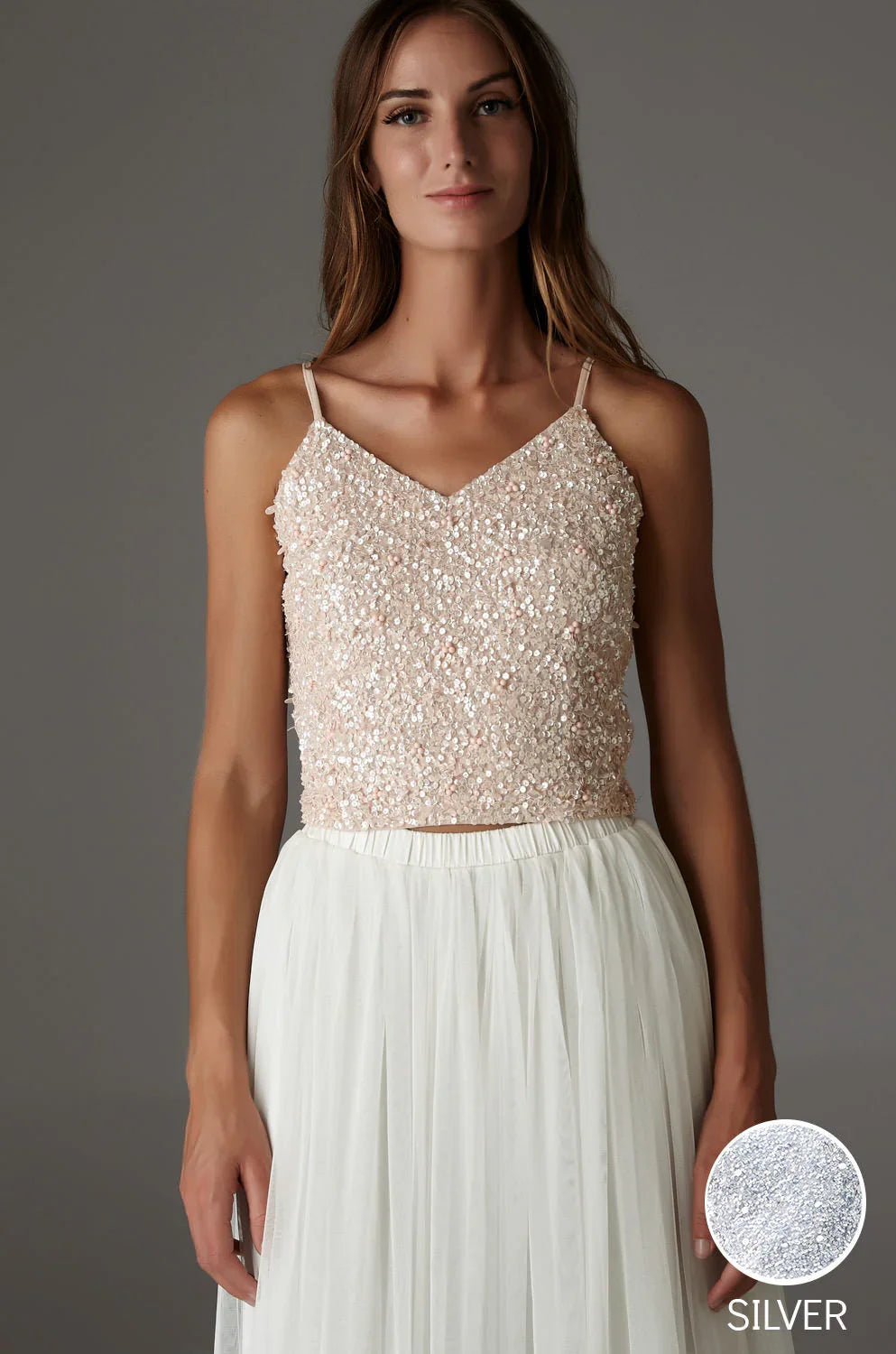 A woman wears the BRIANA TOP SILVER, a sleeveless piece with thin straps and embroidered sequins, paired with a white pleated skirt against a plain gray background. A circular swatch labeled SILVER appears in the bottom right corner.